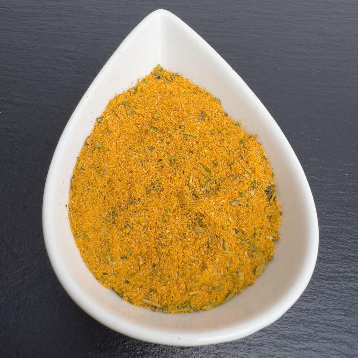 Bratfischwürzer in weißer Schale auf schwarzem Schiefer: orangefarbene Würzmischung mit grünen Kräutern.
