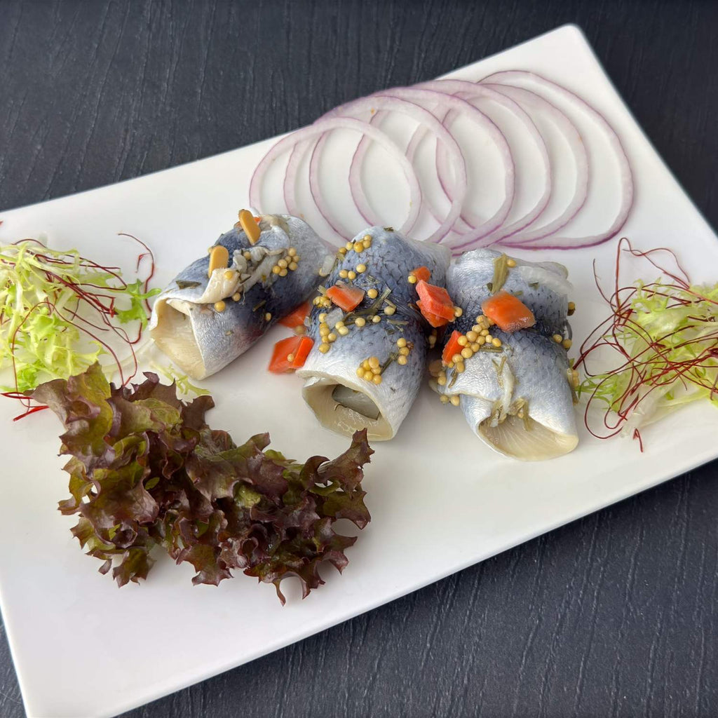 Drei Bremer Gabel-Rollis auf weißer Platte: kleine gerollte Heringsfilets mit Gewürzgurke, Senfsaat und Karotten, daneben Zwiebelringe und Salat als Deko.