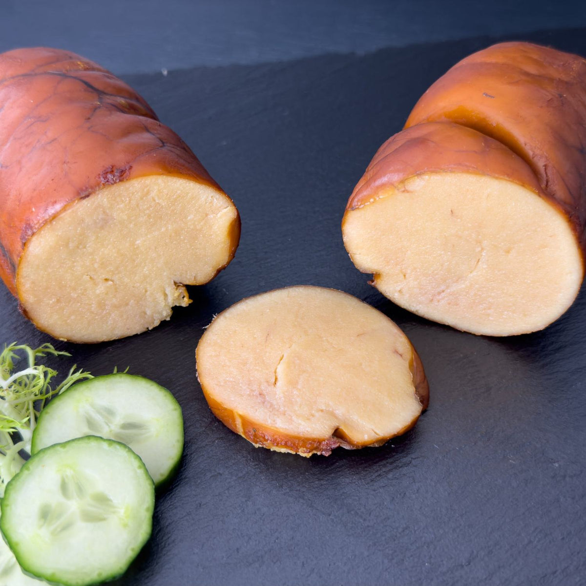 Heißgeräucherter Dorsch-Rogen auf schwarzer Schieferplatte, angeschnitten mit cremig-hellem Inneren und goldbrauner Rogenhaut, daneben Gurke und Salat als Deko.