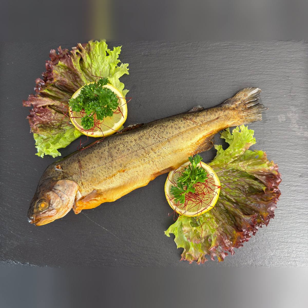   Ganze heißgeräucherte Regenbogenforelle mit goldener Haut auf dunkler Schieferplatte, angerichtet mit Salat, Zitronenscheiben und Petersilie.