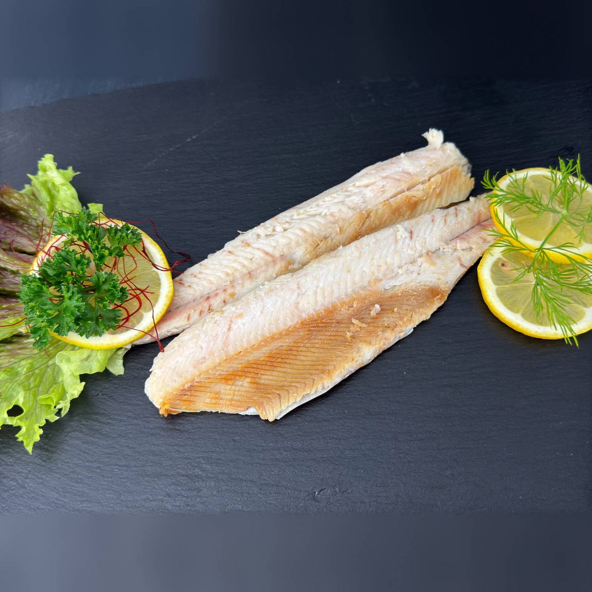 Zwei heißgeräucherte Forellenfilets mit goldenem Rand auf Schieferplatte, angerichtet mit Salat, Zitronenscheiben, Dill und Petersilie.