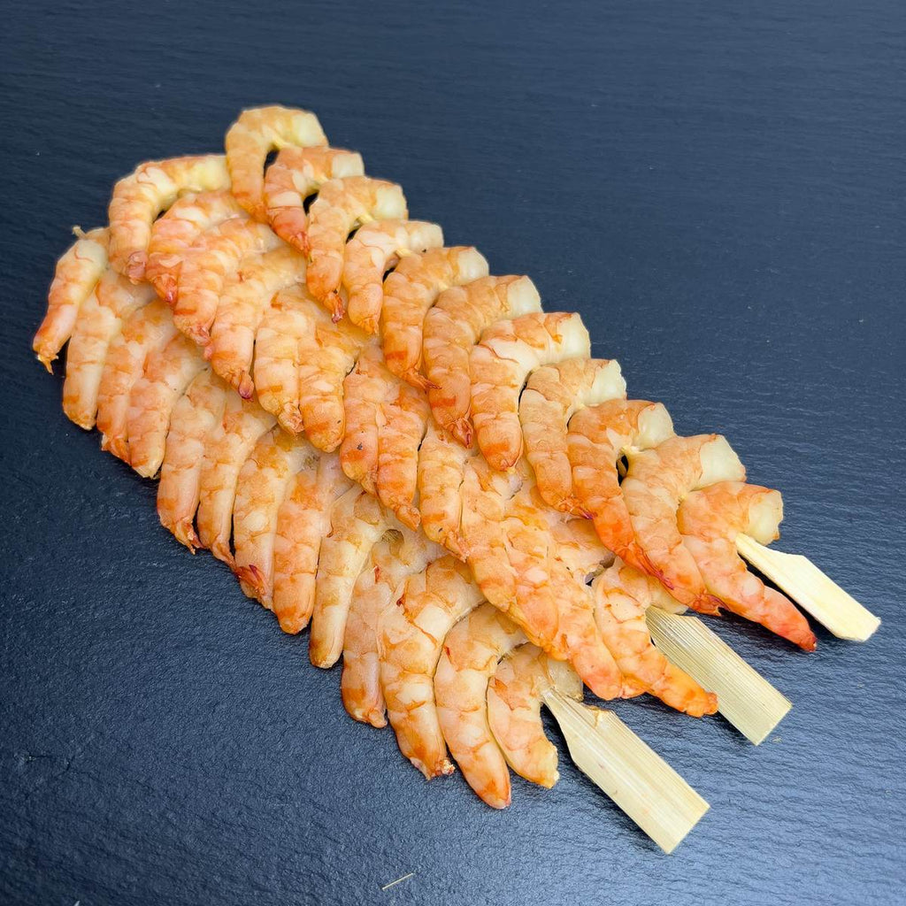 Kaltgeräucherte Black-Tiger-Garnelen auf Bambusspießen, dicht aufgereiht auf dunkler Schieferplatte, servierfertig als Fingerfood.