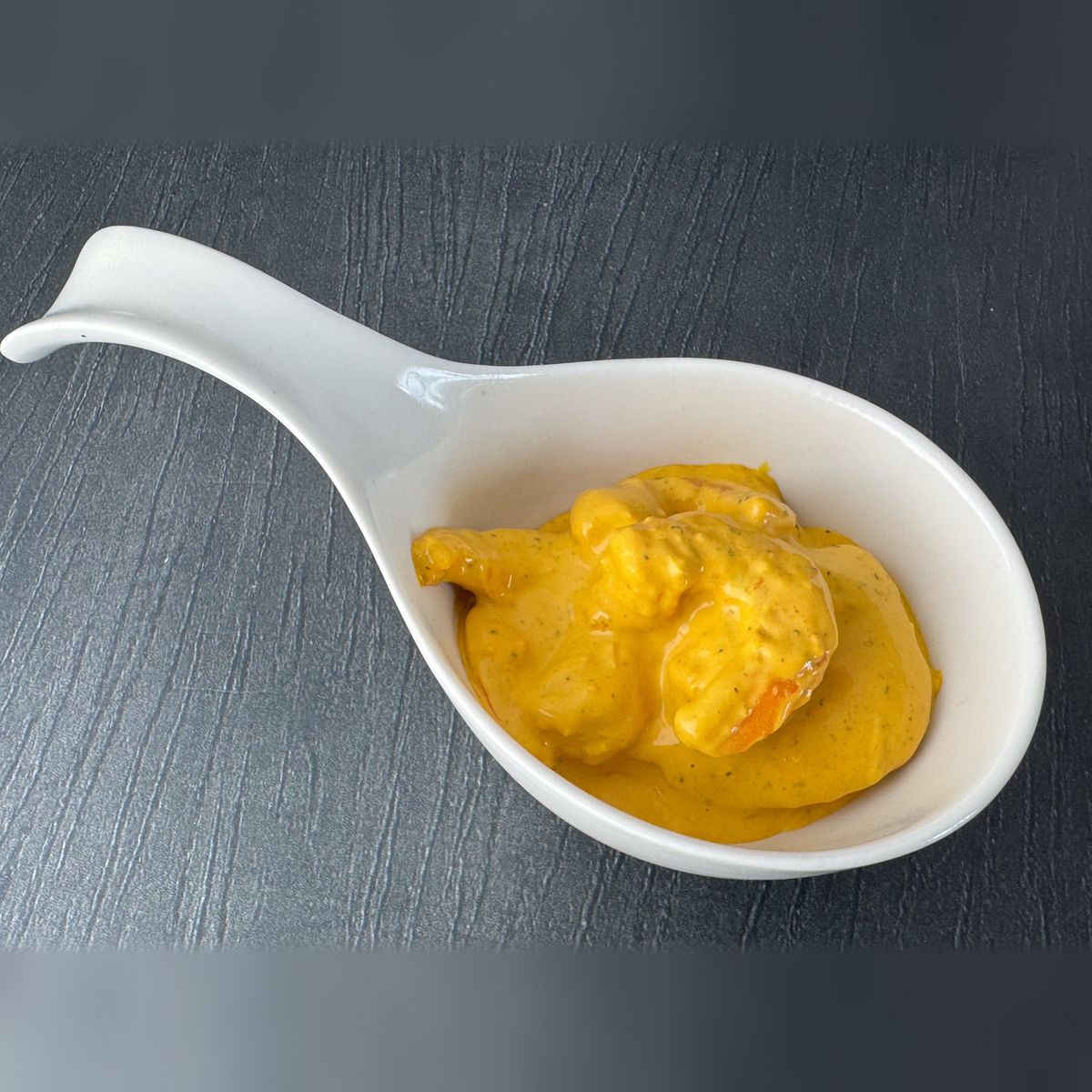 Garnelensalat Bombay in einem weißen Porzellanlöffel auf dunklem Untergrund: Garnelen in cremiger, goldgelber Curry-Mango-Sauce.