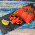 Graved Sockeye-Lachs mit tiefroten Scheiben, Dill und kleinen Lachsrosen mit Queller auf Schieferplatte, daneben eine Zitronenspalte, auf blauem Holz.