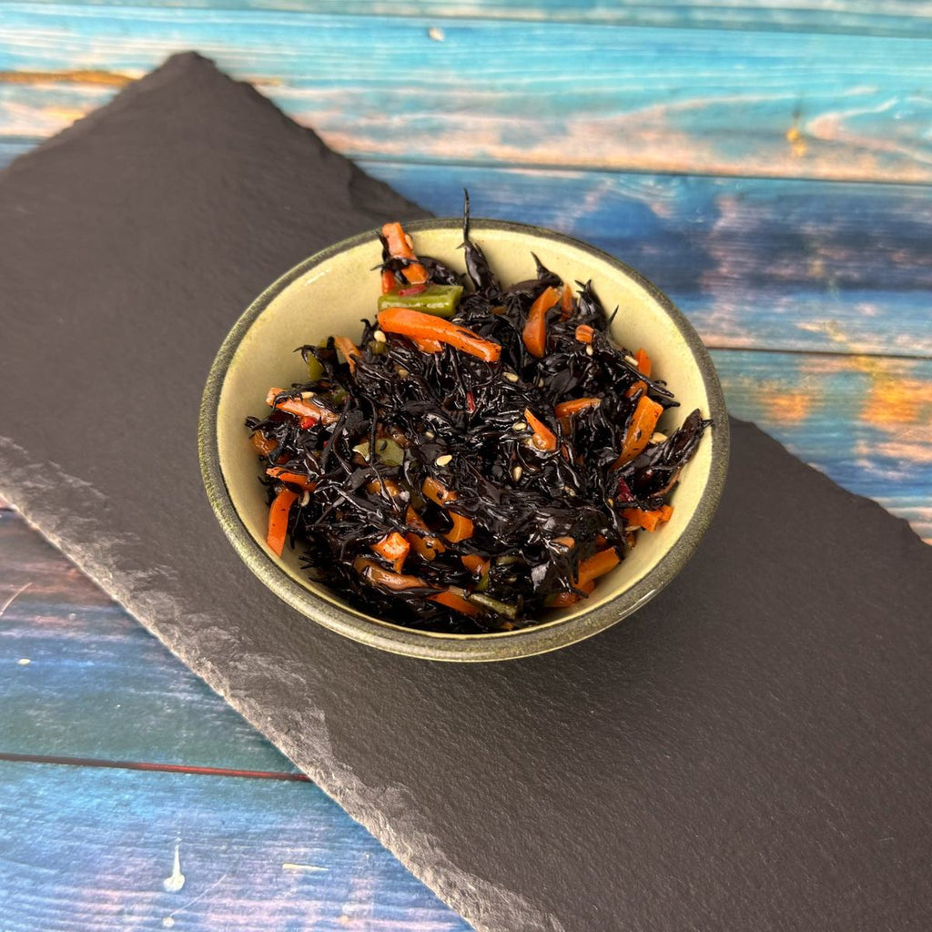 Hijiki-Algensalat in grüner Keramikschale auf Schieferplatte und blauem Holz: schwarze Seealgen mit Karottenstreifen und Sesam.