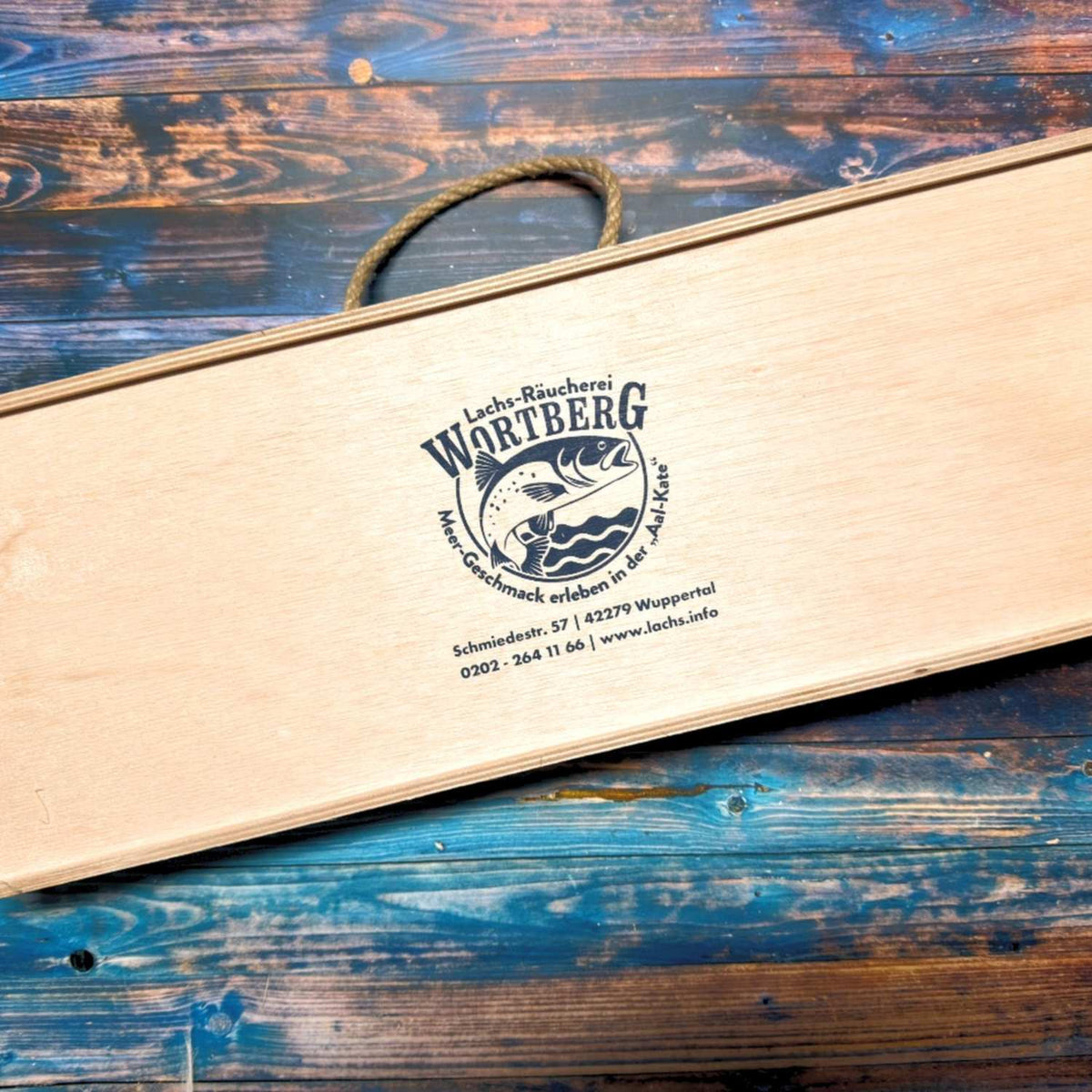 Holzgeschenkbox mit Schiebedeckel, Seilgriff und Logo der Lachs-Räucherei Wortberg auf blauem Holztisch