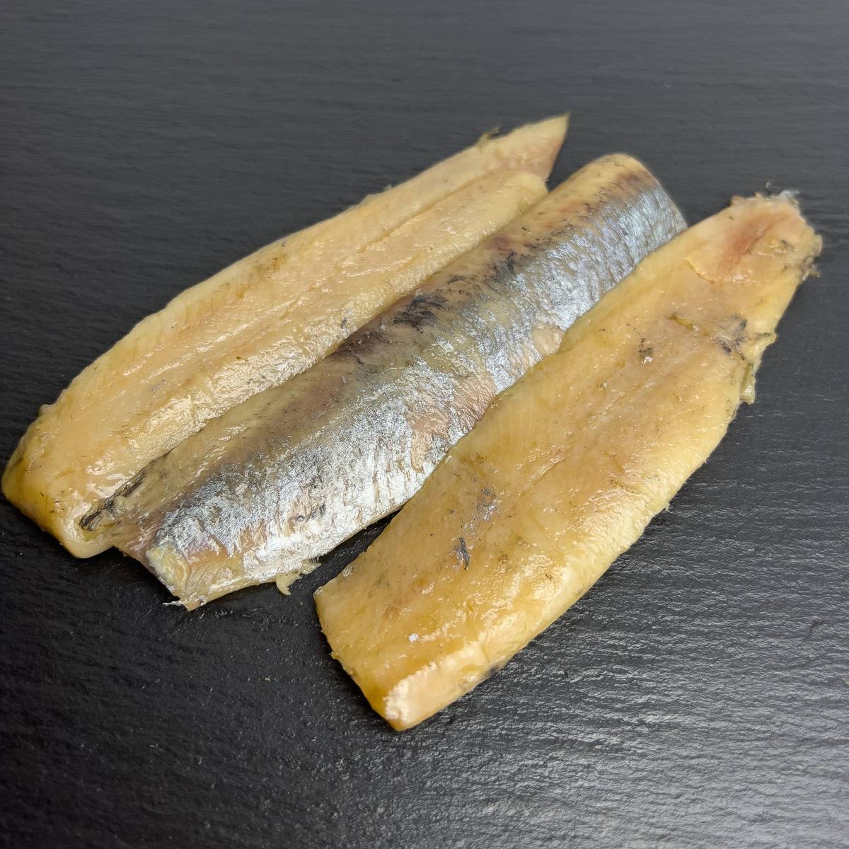 Kaltgeräucherte Matjesfilets auf schwarzem Schiefer, goldfarben mit silbriger Haut.