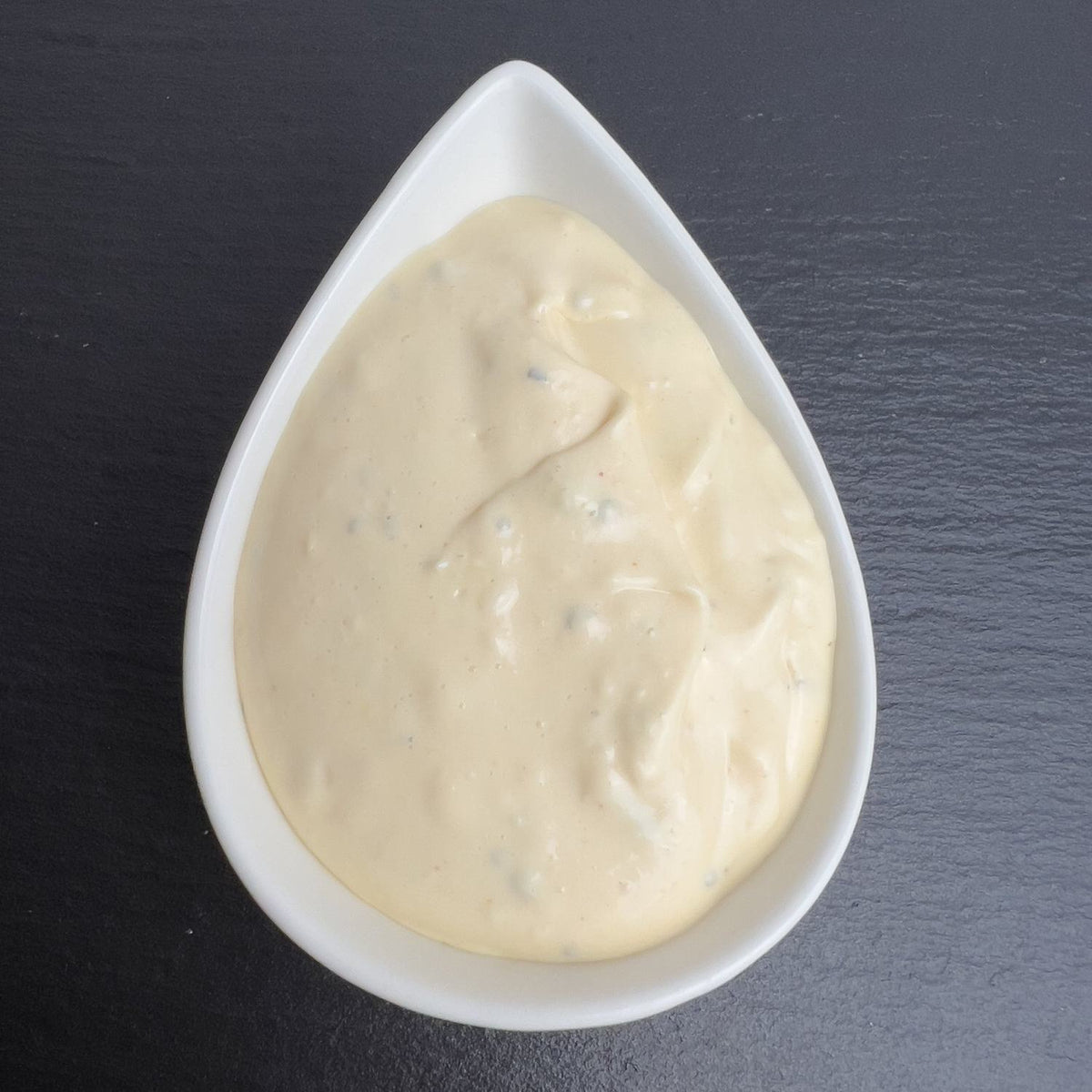 Knoblauch-Dip in weißer Schale auf schwarzem Schiefer: cremiger heller Dip mit Kräutern.