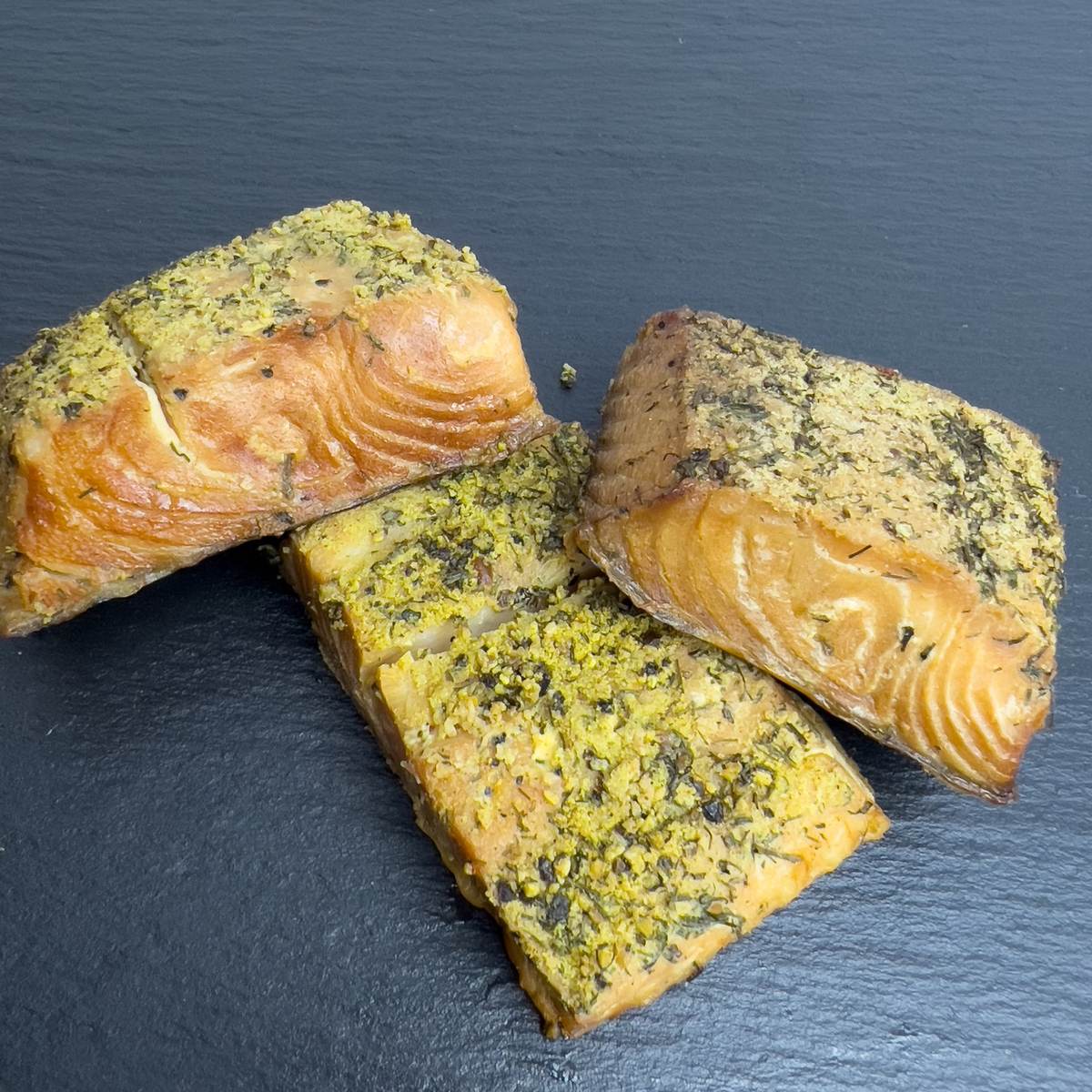 Heißgeräucherte Stücke vom Schwarzen Heilbutt mit grüngelber Kräuterkruste aus Dill und Gewürzen auf dunkler Schieferplatte.
