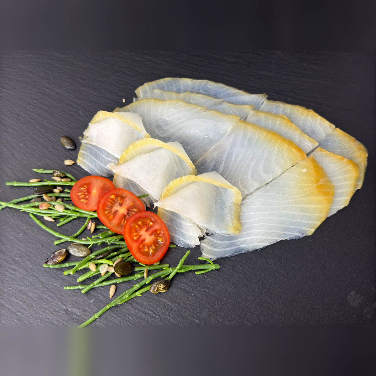 Hauchdünne Scheiben vom kaltgeräucherten Schwarzen Heilbutt mit goldenem Rand auf Schieferplatte, dekoriert mit Tomate und Queller.