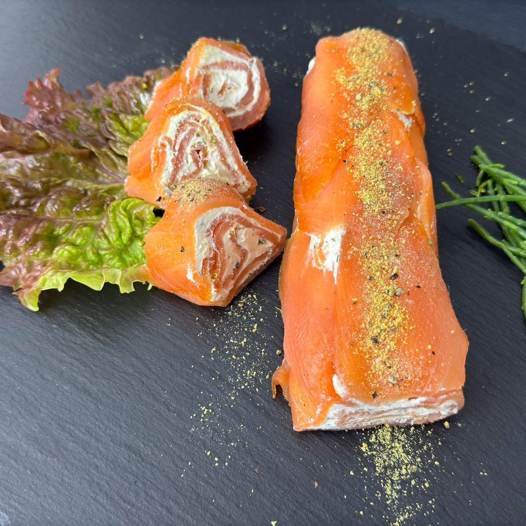 Lachs-Rolle mit Frischkäse und Wasabi auf schwarzer Schieferplatte, im Anschnitt mit Frischkäse-Spirale, daneben Salat und Queller als Deko.
