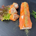 Lachs-Rolle mit Frischkäse und Wasabi auf schwarzer Schieferplatte, im Anschnitt mit Frischkäse-Spirale, daneben Salat und Queller als Deko.