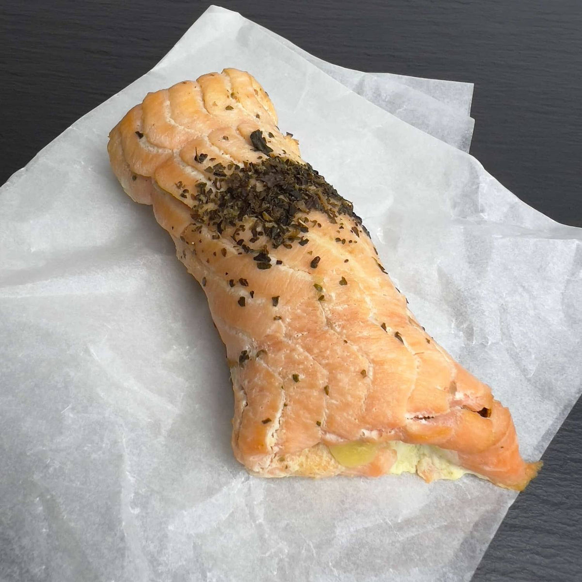 Heißgeräucherte Lachs-Roulade auf Pergamentpapier, bestreut mit Kräutern, an der Schnittseite ist die Füllung aus Pesto und Mozzarella zu sehen.