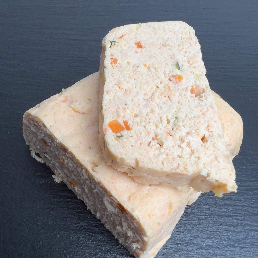 Hausgemachte Lachs-Terrine in Scheiben auf schwarzer Schieferplatte, zartrosa und schnittfest, mit bunten Möhren- und Zucchiniwürfeln.