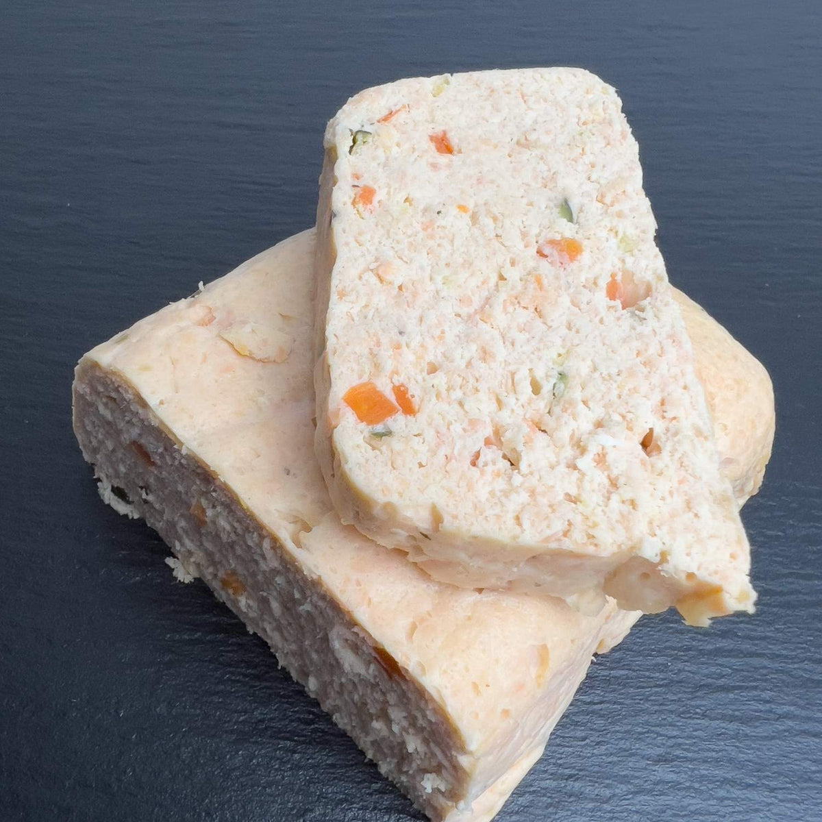 Hausgemachte Lachs-Terrine in Scheiben auf schwarzer Schieferplatte, zartrosa und schnittfest, mit bunten Möhren- und Zucchiniwürfeln.