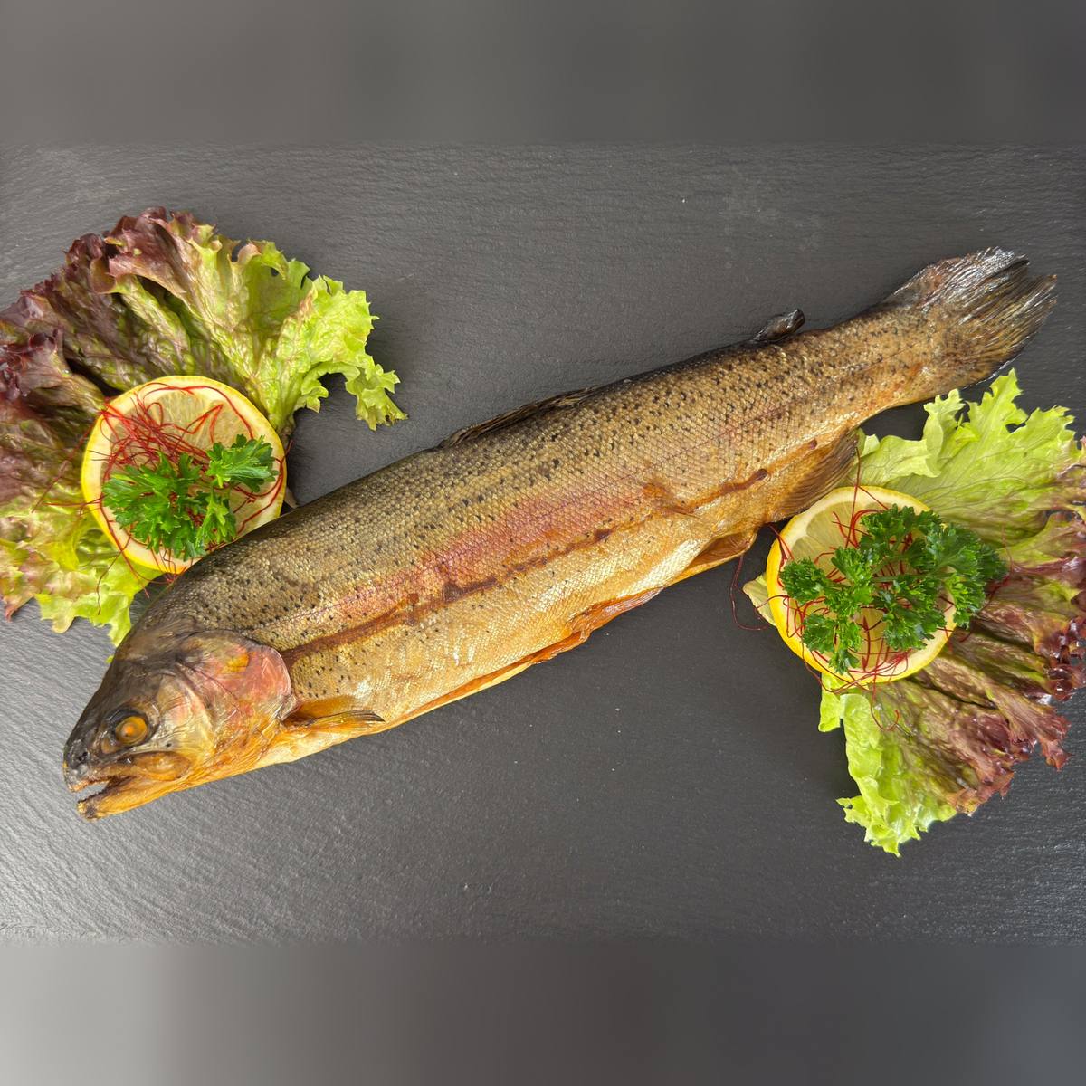   Ganze heißgeräucherte Lachsforelle mit rosa schimmernder, goldbrauner Haut auf Schieferplatte, garniert mit Salat, Zitrone und Petersilie.