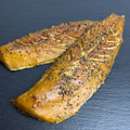 Zwei heißgeräucherte Makrelenfilets mit goldbrauner Oberfläche und grober Pfefferkruste auf dunkler Schieferplatte.