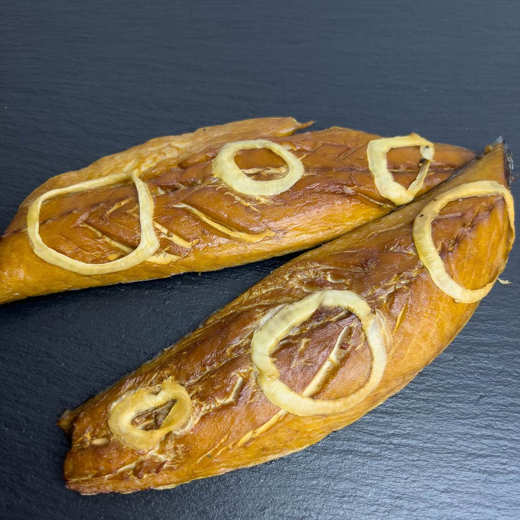 Zwei heißgeräucherte Makrelenfilets mit goldbrauner Haut und weichen Zwiebelringen auf dunkler Schieferplatte.