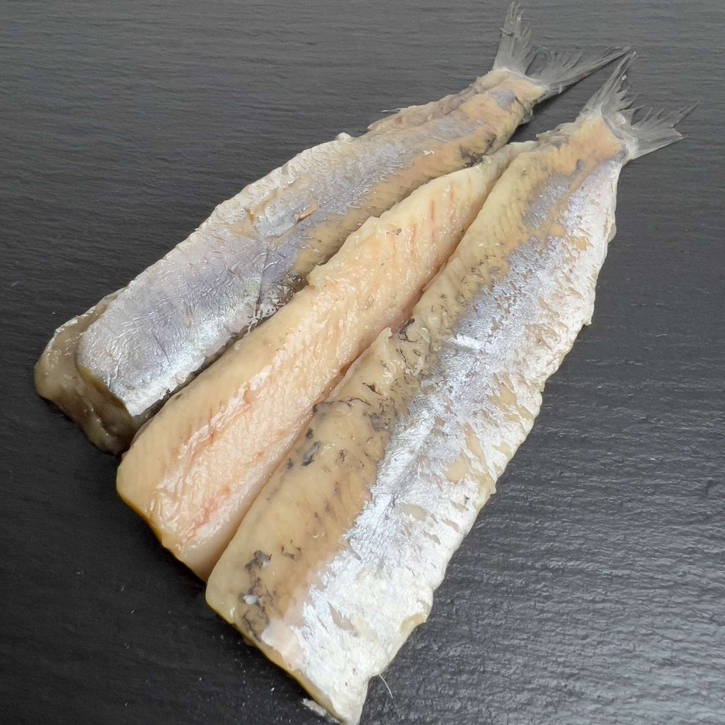 Matjes-Doppelfilets auf schwarzem Schiefer: silbrig glänzende Heringsfilets, an der Schwanzflosse verbunden.