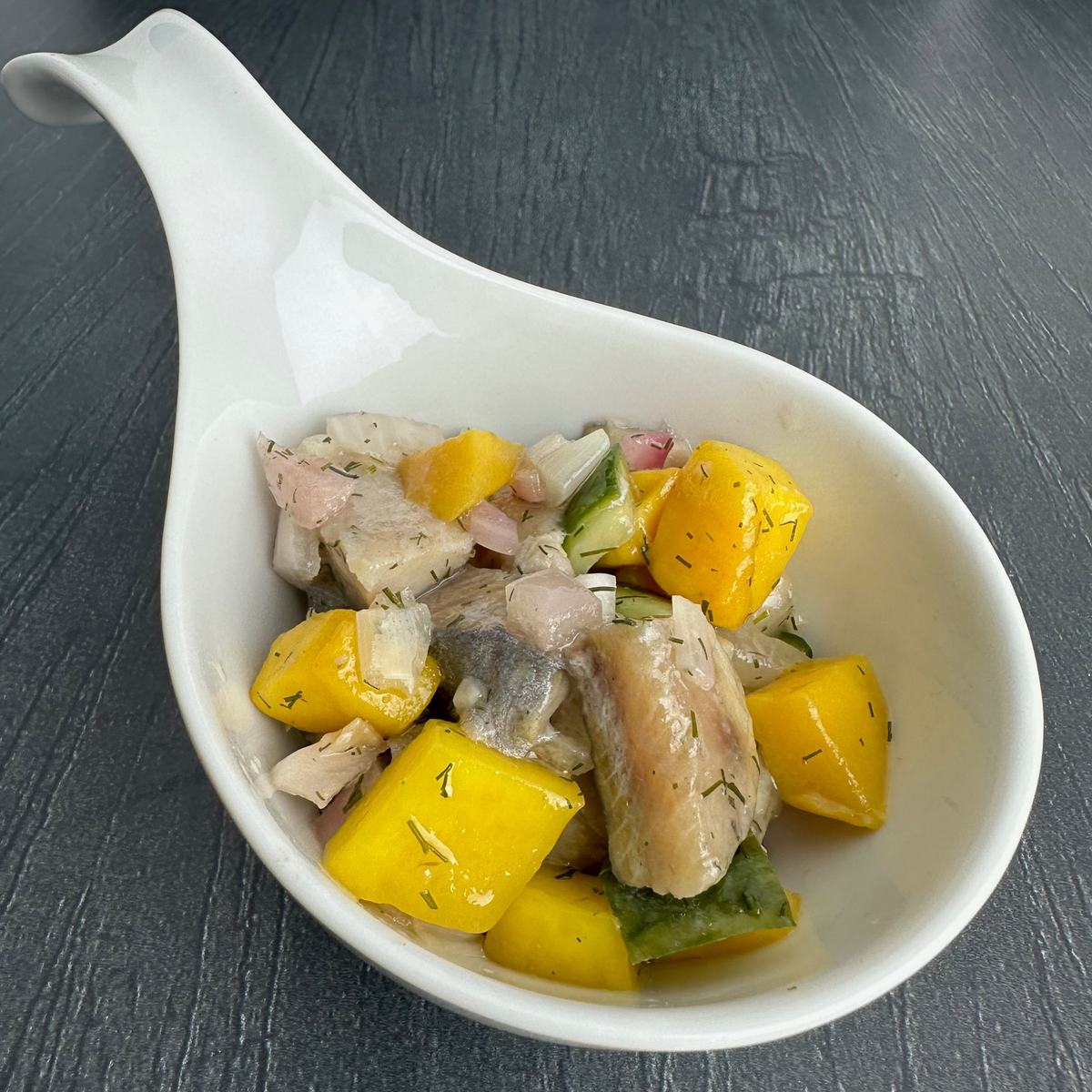 Hausgemachter Matjes-Mango-Salat in einem weißen Porzellanlöffel: Matjesstücke mit gelben Mangowürfeln, Gurke, roten Zwiebeln und frischem Dill.