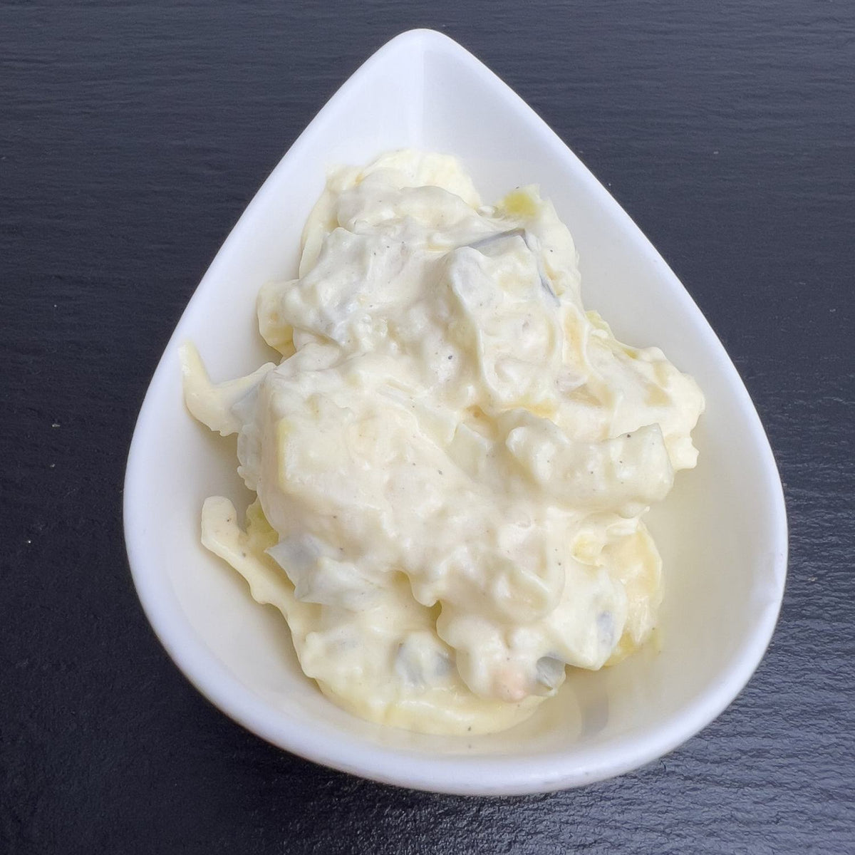 Pell-Kartoffelsalat mit Ei und Gewürzgurke in cremigem Mayonnaise-Dressing, in weißer Schale auf schwarzem Schiefer.