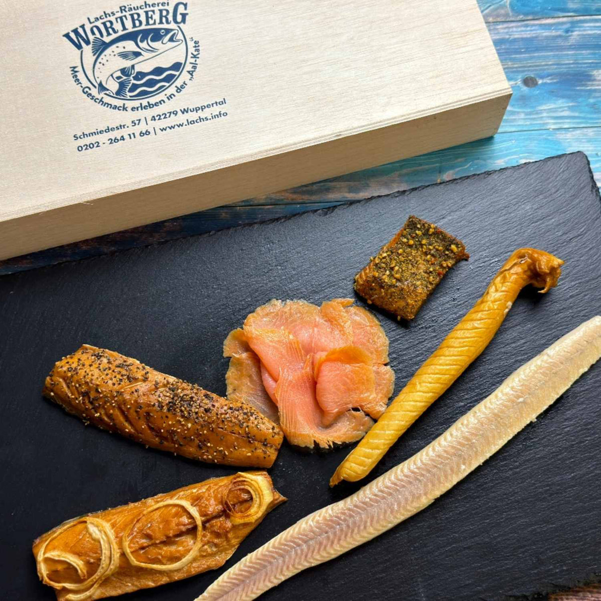 Präsent Fischers Feinste mit Aalfilet, Makrelenfilet mit Pfeffer, Schillerlocke, Kräuter-Butt und Räucherlachs auf einer Schieferplatte vor der Holzbox