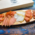 Präsent Lachs trifft Butt: fünf Sorten Räucherlachs und Heilbutt in Scheiben auf einem Holzbrett vor der Holzgeschenkbox
