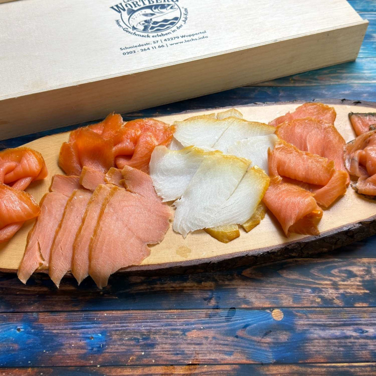Präsent Lachs trifft Butt: fünf Sorten Räucherlachs und Heilbutt in Scheiben auf einem Holzbrett vor der Holzgeschenkbox