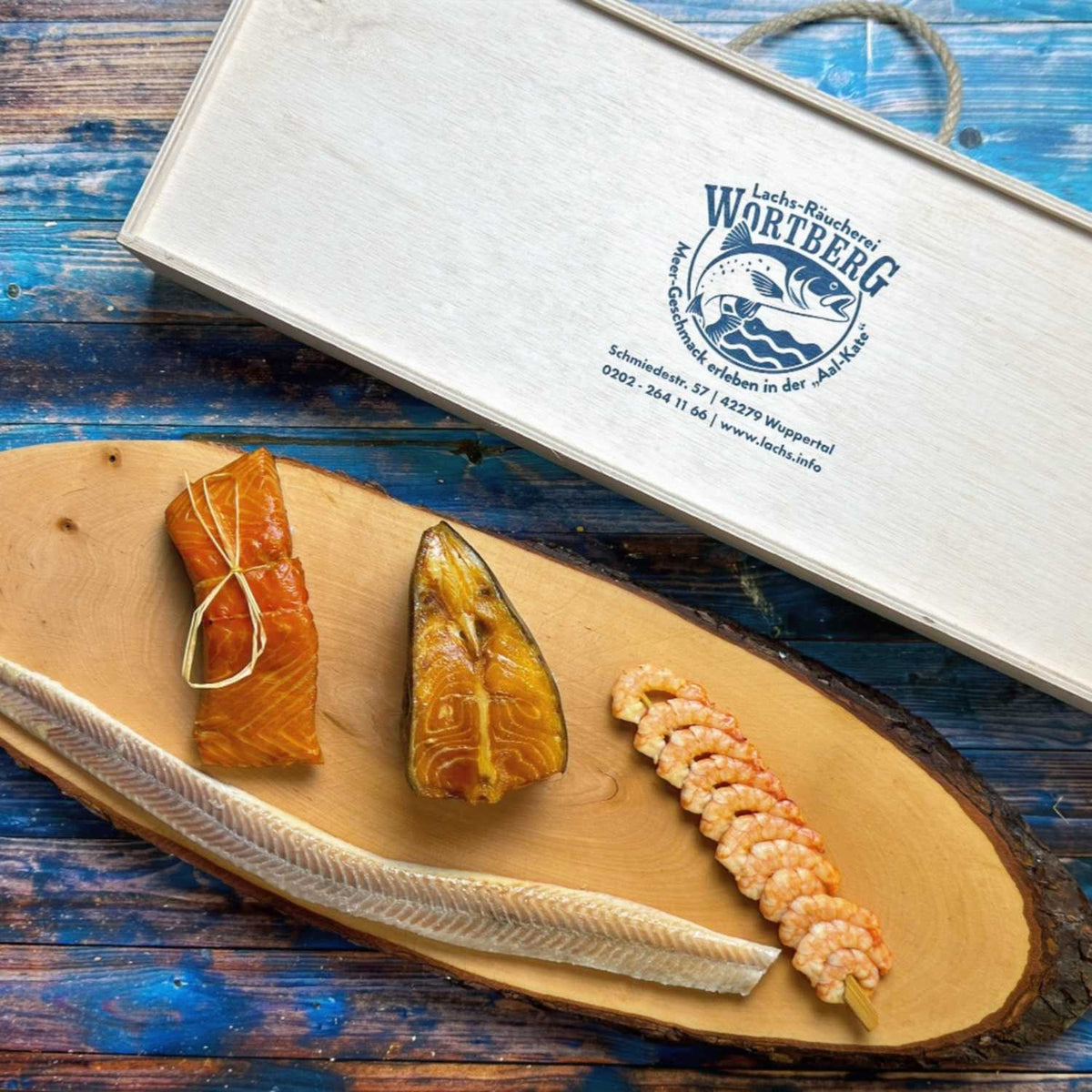Präsent Meer für Feinschmecker mit Striemenlachs, Heilbutt, Garnelen-Spieß und Aalfilet auf einem Holzbrett vor der Holzbox