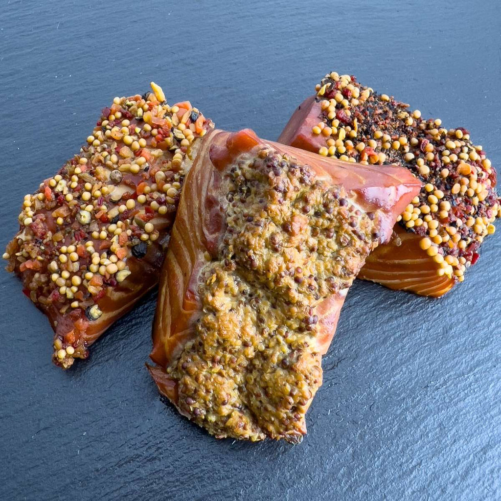 Drei Räucherlachs-Pralinen auf Schieferplatte: heißgeräucherte Lachsstücke mit Würzkrusten aus Paprika-Pfeffer, Dijon-Senf und Pfeffer-Senf.
