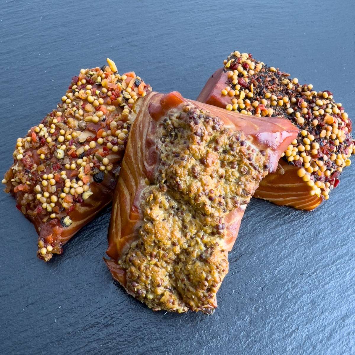 Drei Räucherlachs-Pralinen auf Schieferplatte: heißgeräucherte Lachsstücke mit Würzkrusten aus Paprika-Pfeffer, Dijon-Senf und Pfeffer-Senf.