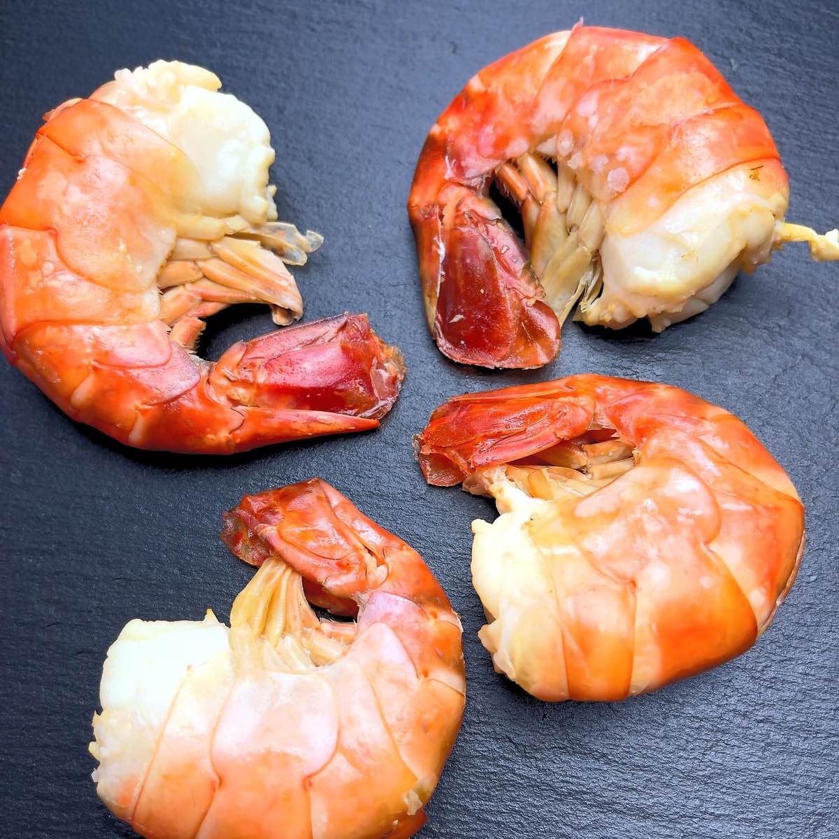Vier große kaltgeräucherte Riesengarnelen-Schwänze mit orange-roter Schale auf dunkler Schieferplatte, saftiges weißes Fleisch sichtbar.