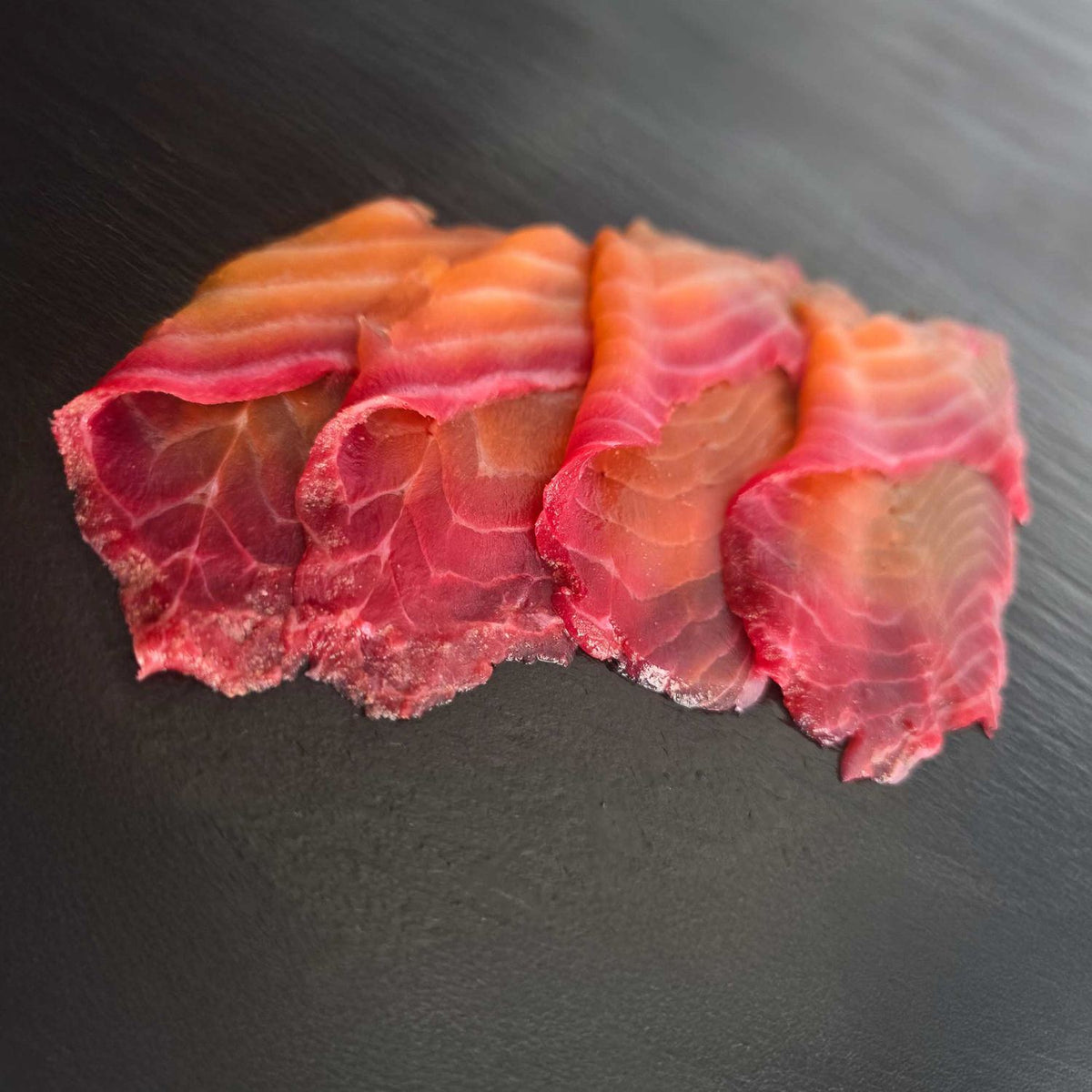 Hauchdünne Scheiben Rote-Bete-Lachs auf dunkler Schieferplatte, außen kräftig rot, innen zartrosa, nach Graved-Art gebeizt.