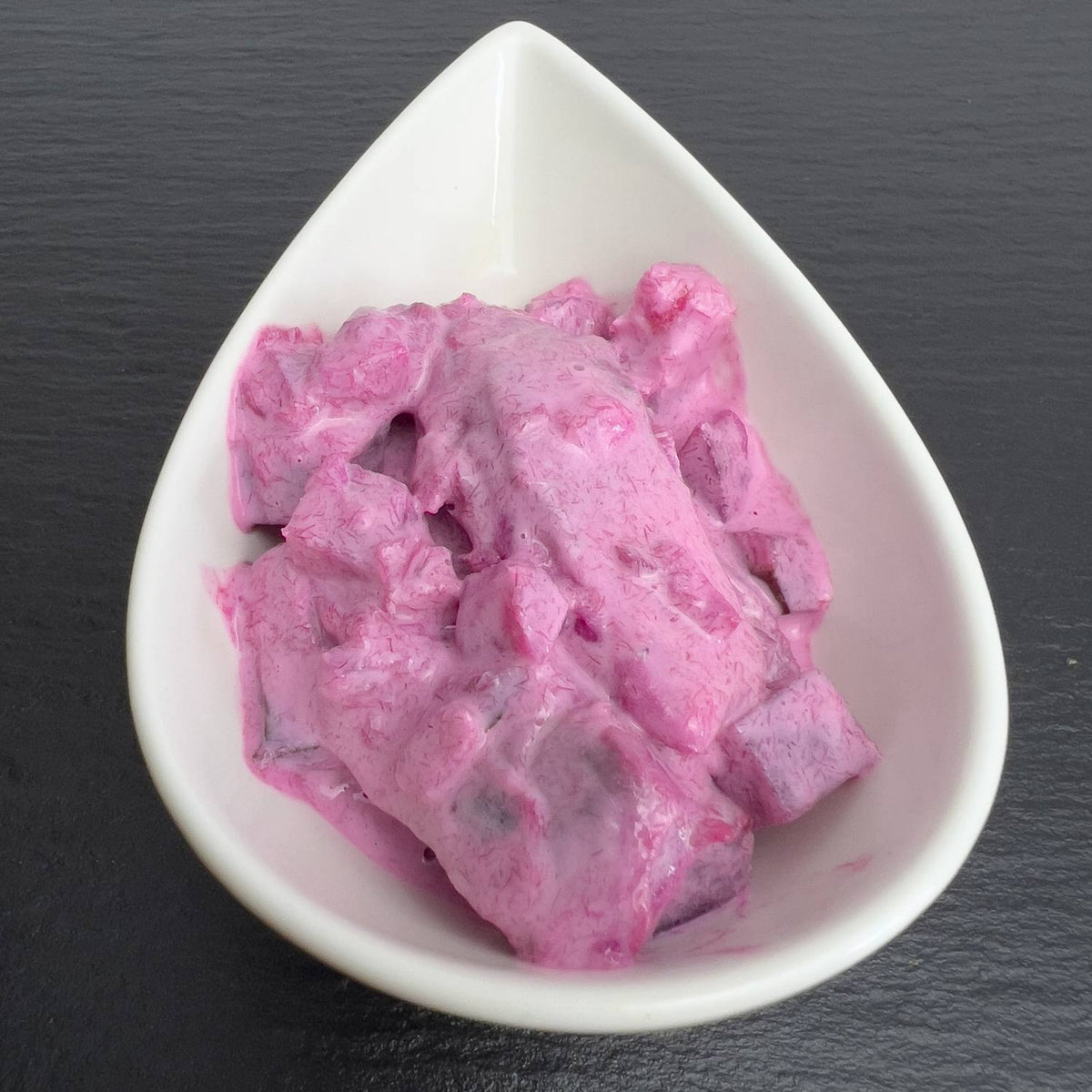 Roter Heringssalat in weißer Schale auf schwarzem Schiefer: Hering mit Roter Bete, Apfel und Gewürzgurke in leuchtend roter, cremiger Sauce.