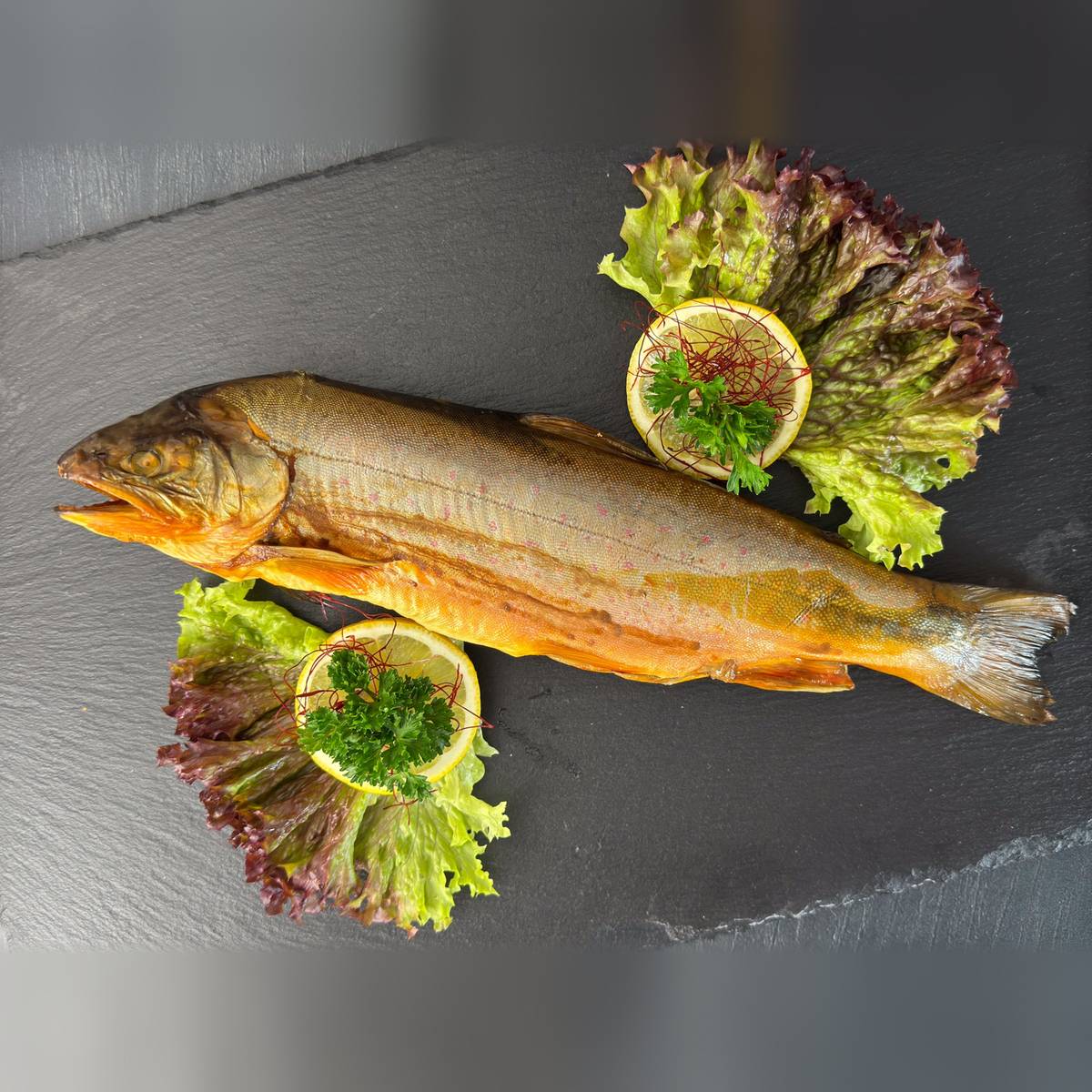   Ganzer heißgeräucherter Saibling mit goldorangener Haut und rosa Punkten auf Schieferplatte, angerichtet mit Salat, Zitrone und Petersilie.