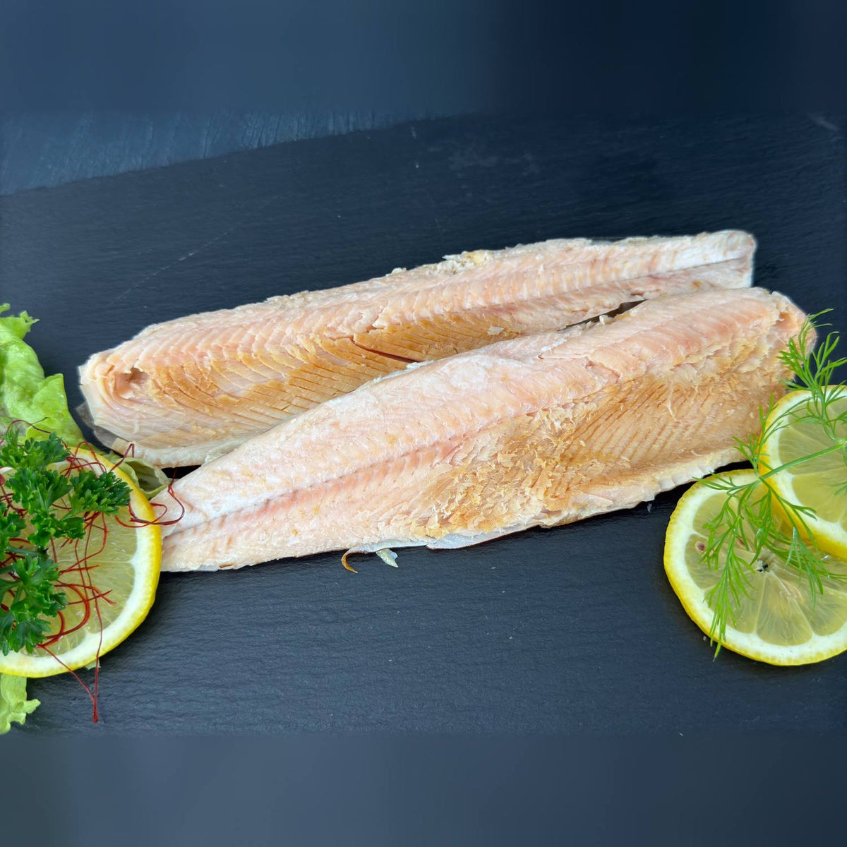Zwei heißgeräucherte Saiblingsfilets mit rosigem Fleisch und goldenem Rand auf Schieferplatte, garniert mit Salat, Zitrone, Dill und Petersilie.