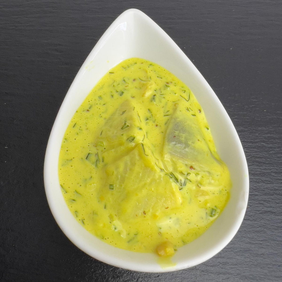 Senf-Kräuter-Happen in weißer Schale auf schwarzem Schiefer: Heringsstücke in gelber Honig-Senf-Sauce mit frischen Kräutern.