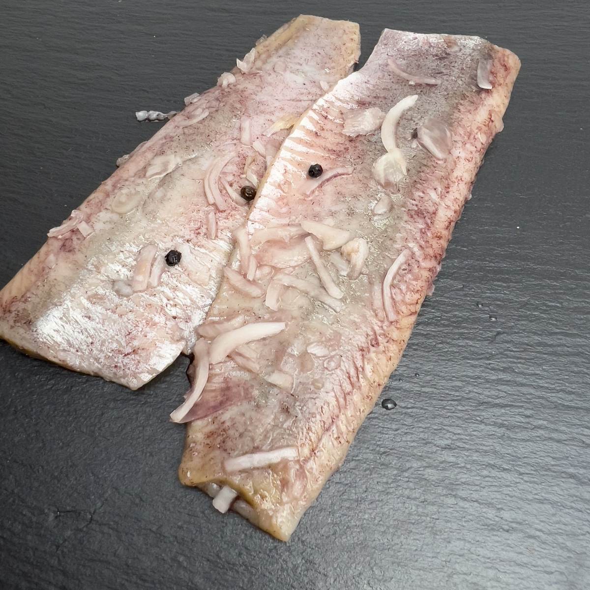 Sherry-Matjesfilets auf schwarzem Schiefer, rosa marmoriert mit Zwiebelstreifen und Pfefferkörnern.