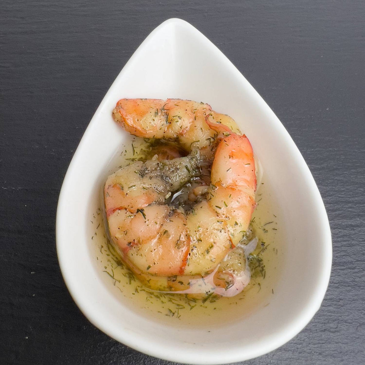 Black-Tiger-Garnelen in Knoblauch-Kräuter-Öl mit Dill in weißer Schale auf schwarzem Schiefer.
