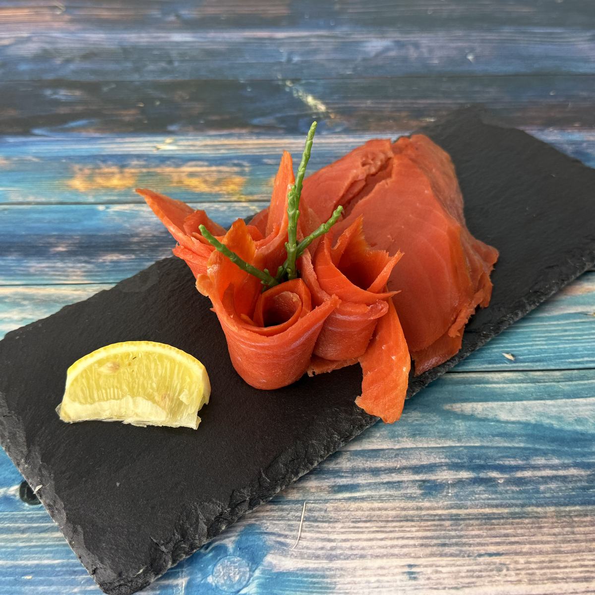 Sockeye-Wildräucherlachs als tiefrote Lachsrosen mit Queller und Scheiben auf Schieferplatte, daneben eine Zitronenspalte, auf blauem Holz angerichtet.