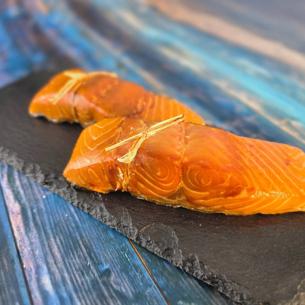 Zwei heißgeräucherte Lachsfilets, von Hand mit Bast gebunden, leuchtend orange auf Schieferplatte vor blauem Holz.