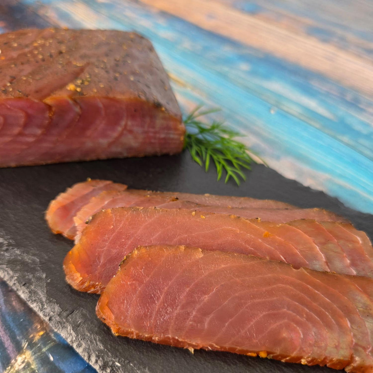 Thunfischfilet Pastrami auf Schieferplatte: ein Stück mit würziger Pfefferkruste, davor zartrote, kaltgeräucherte Scheiben und ein Zweig frischer Dill.