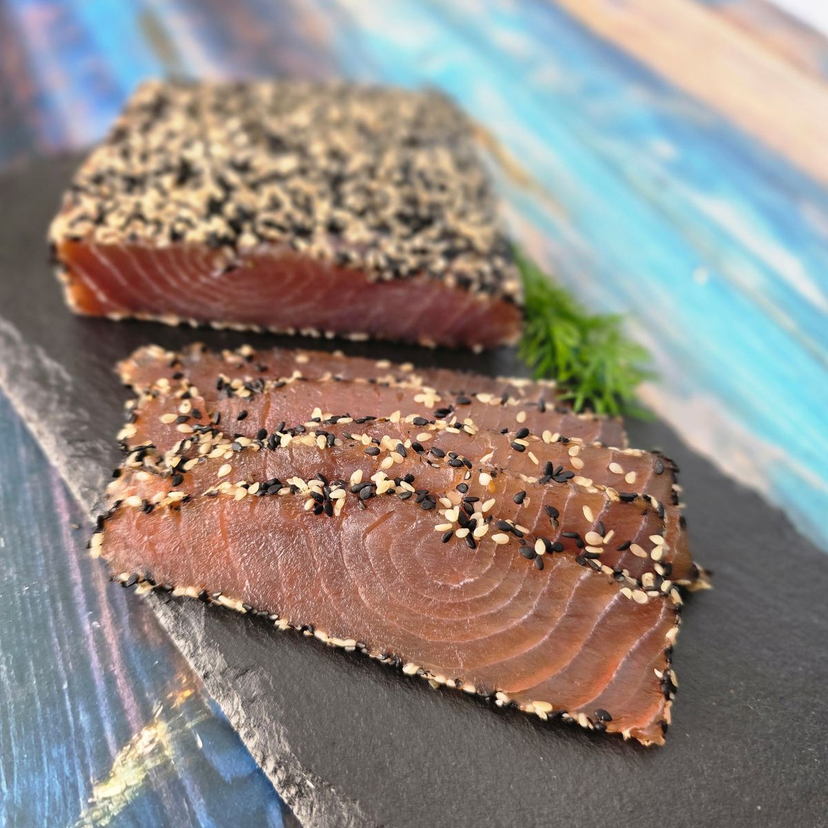 Thunfischfilet Sesam am Stück und in Scheiben auf Schieferplatte: tiefrotes, kaltgeräuchertes Thunfischfilet mit schwarz-weißem Sesammantel und frischem Dill.