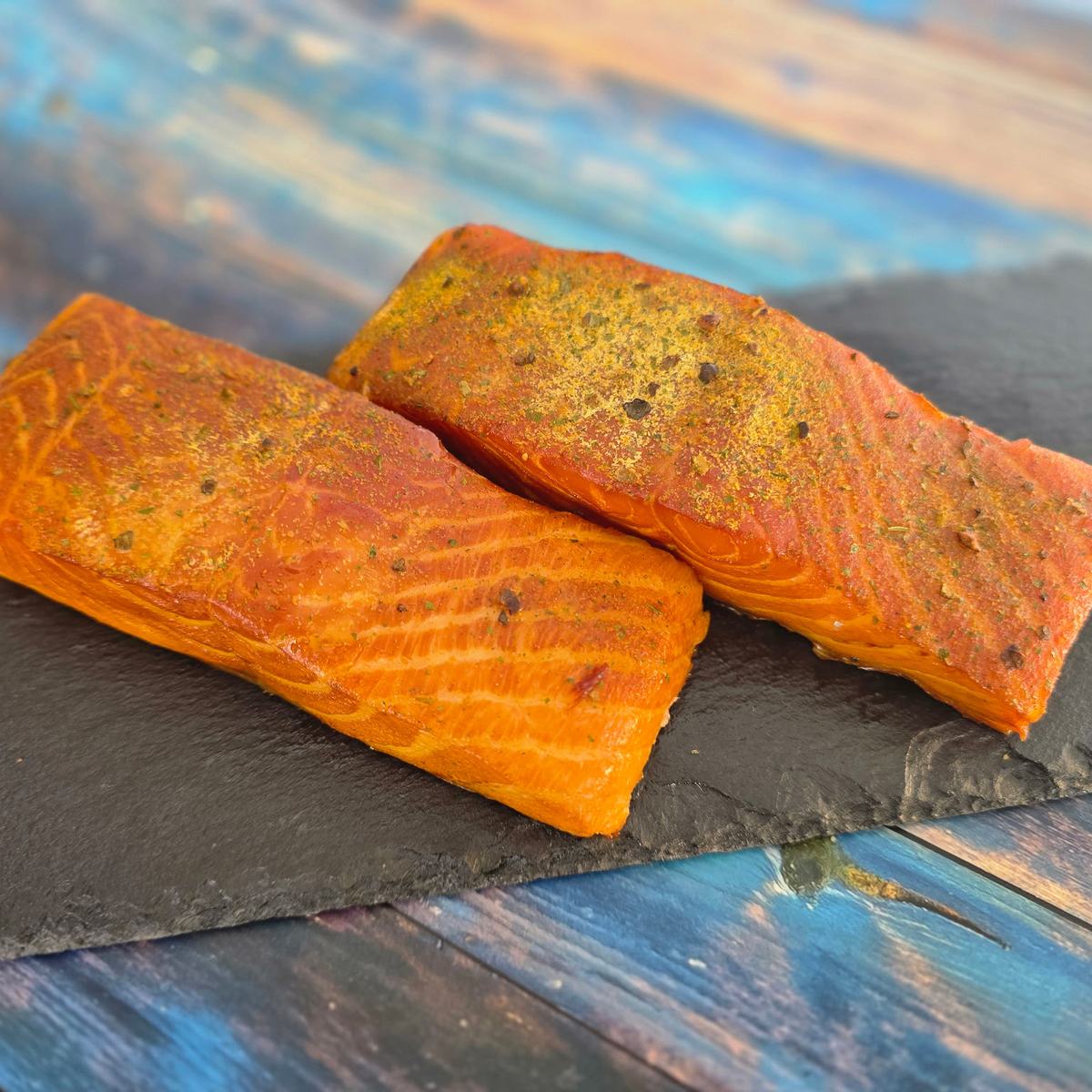   Zwei heißgeräucherte Lachsfilets mit würziger Kruste aus Wacholder und Zitronenpfeffer auf Schieferplatte vor blauem Holz.